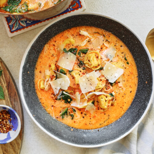 creamy tuscan tortellini soup in a black speckled bowl with a gold spoon beside it