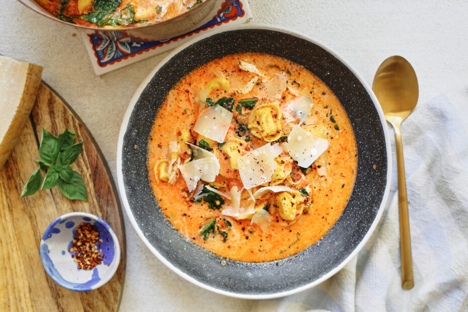 creamy tuscan tortellini soup in a black speckled bowl with a gold spoon beside it