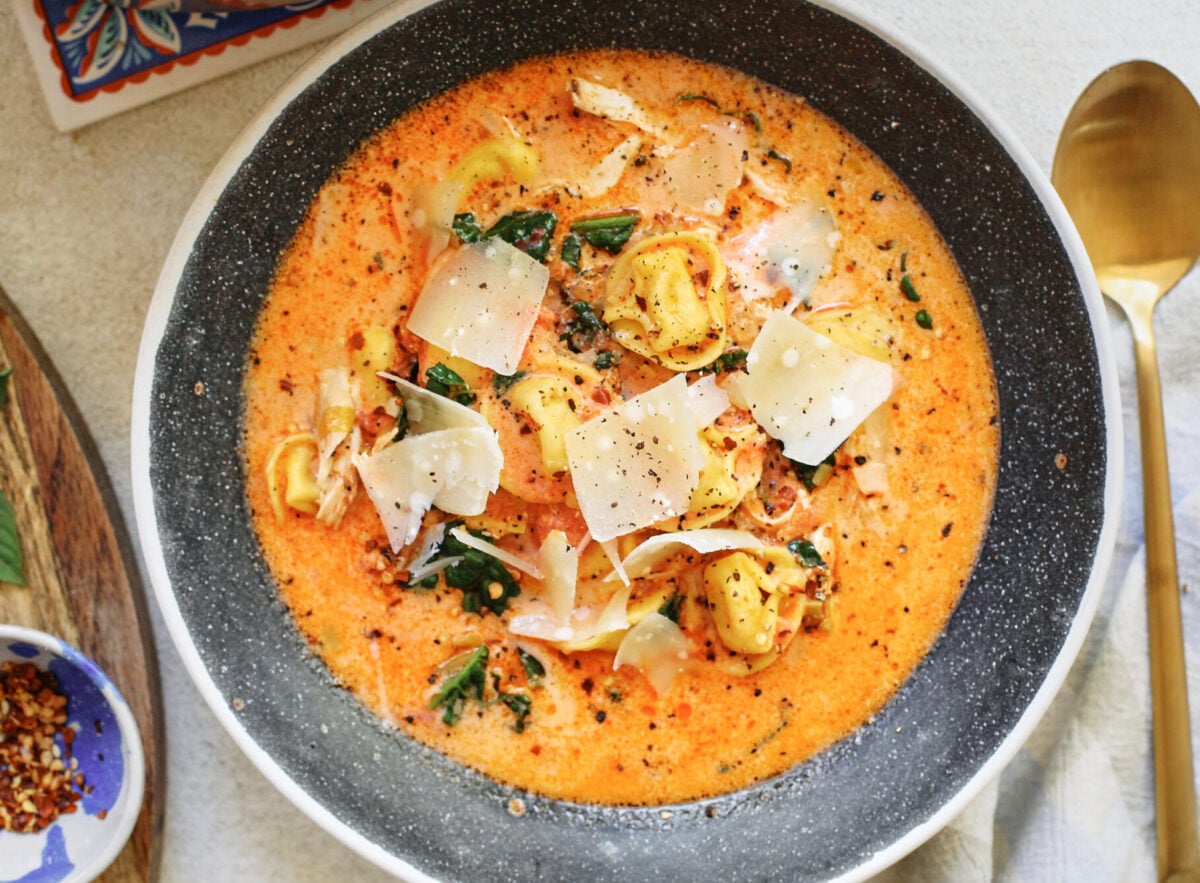 creamy tuscan tortellini soup in a black speckled bowl with a gold spoon beside it