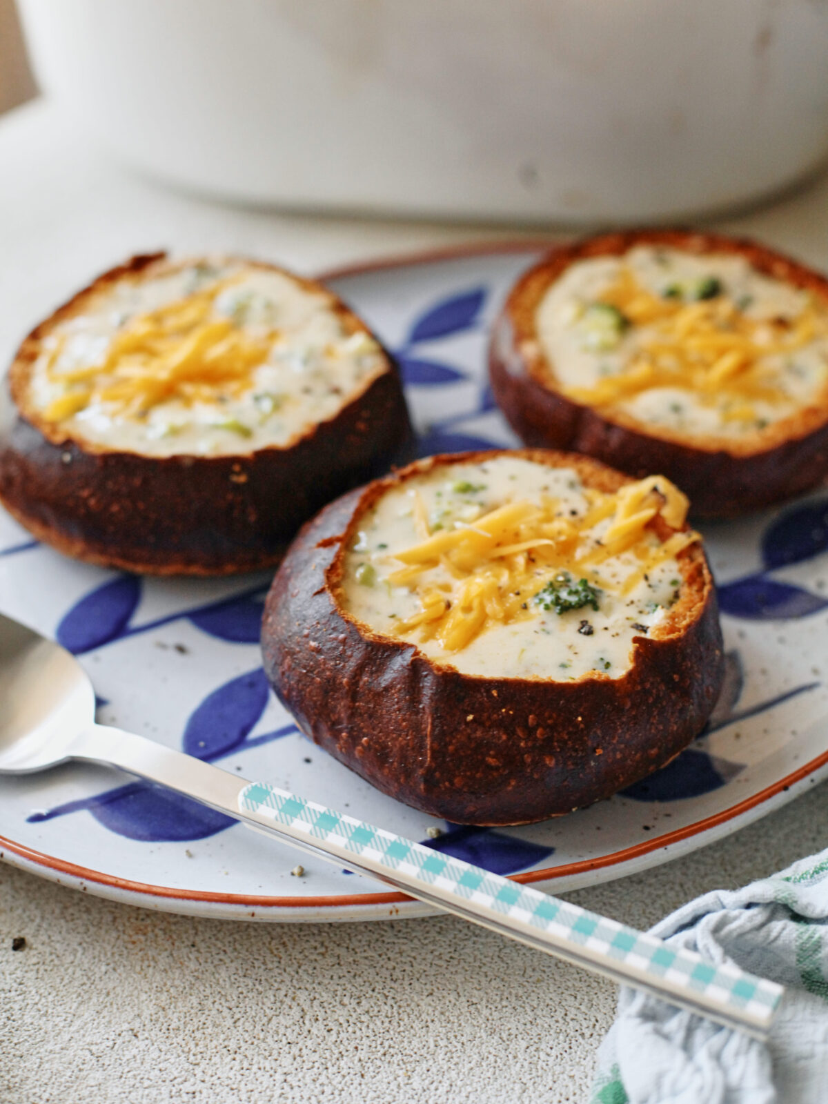 three pretzel bun bread bowls filled with broccoli cheddar soup on a white platter with blue leaf motifs