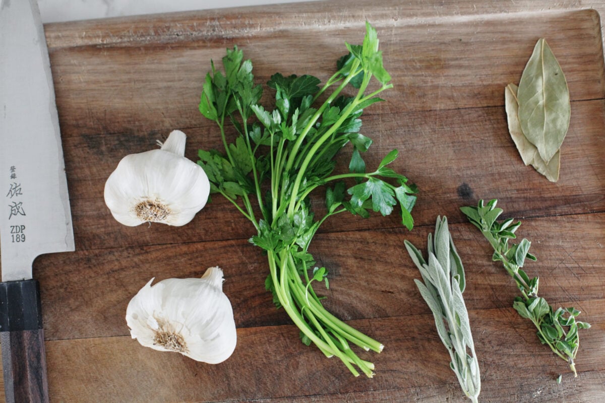 two heads of garlic a bunch of parsley, sage leaves, thyme, and bay leaves on a wooden cutting board