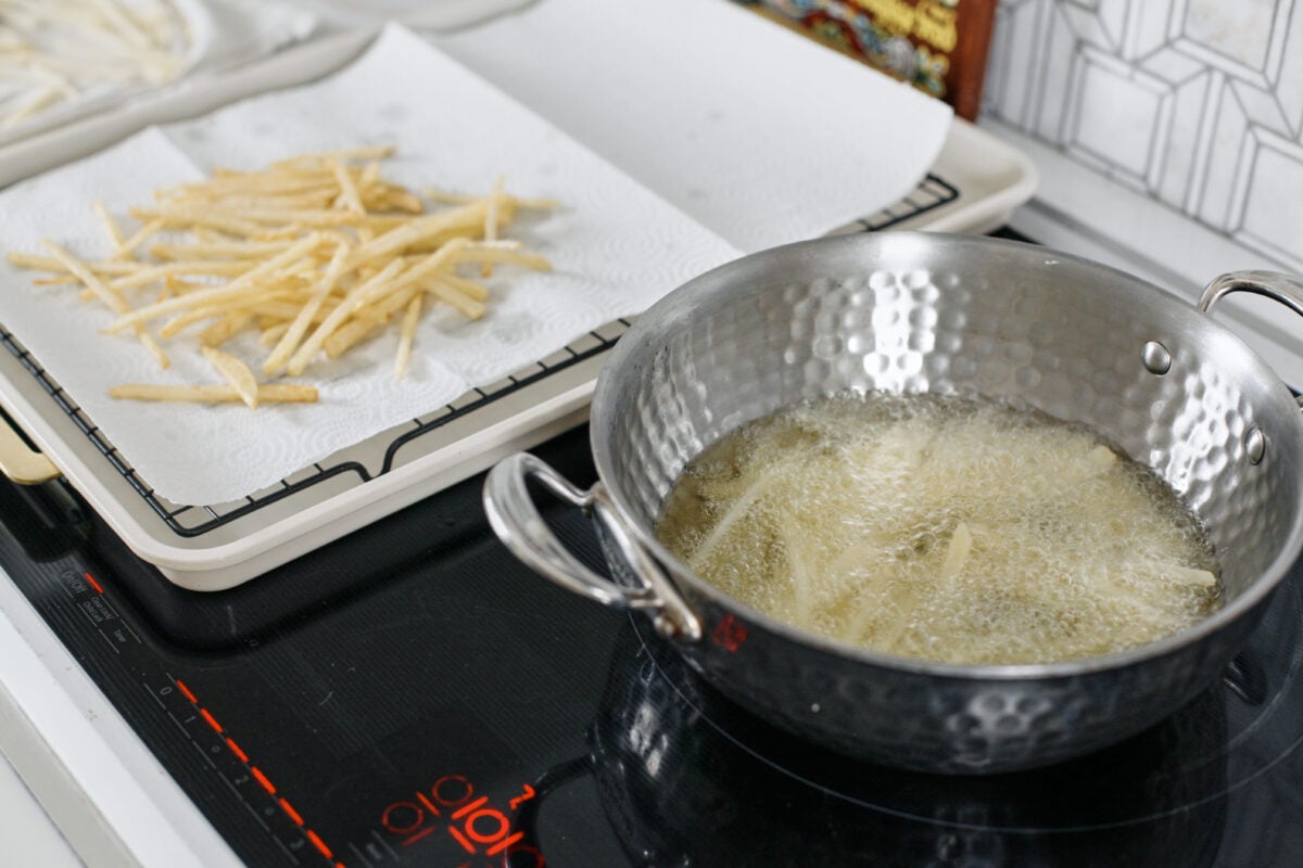 french fries frying in a shallow pot of oil, with a batch of cooked fries in the background
